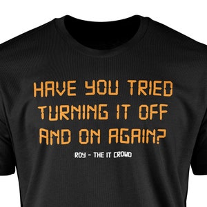 May include: Black t-shirt featuring the orange text "HAVE YOU TRIED TURNING IT OFF AND ON AGAIN?" and "ROY - THE IT CROWD". The shirt has a crew neck.