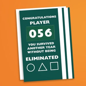 May include: A greeting card with a dark green background and white text that reads "CONGRATULATIONS PLAYER 056 YOU SURVIVED ANOTHER YEAR WITHOUT BEING ELIMINATED" with a circle, triangle, and square symbol. The card is on a white background.
