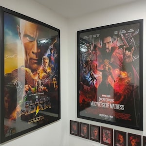 May include: Two framed film posters are displayed on a white wall. One is for "Black Adam", featuring Dwayne Johnson, and the other is for "Doctor Strange in the Multiverse of Madness". The "Doctor Strange" poster has a red and purple color scheme.