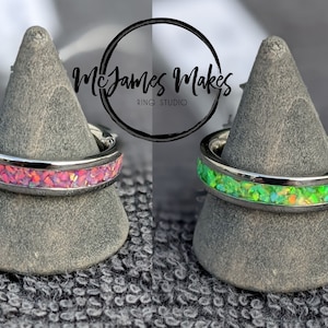 May include: Two stainless steel rings with a thin band of opal inlay. The ring on the left has pink opal inlay and the ring on the right has green opal inlay. McJames Makes Ring Studio is printed on the background.