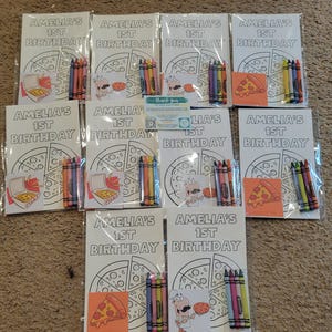 K&ouml;nnte beinhalten: Eine Sammlung von Malb&uuml;chern und Buntstiften f&uuml;r den ersten Geburtstag eines Kindes. Jedes Buch zeigt ein Pizza-Design und den Text "AMELIA'S 1ST BIRTHDAY". Buntstifte in verschiedenen Farben sind enthalten.