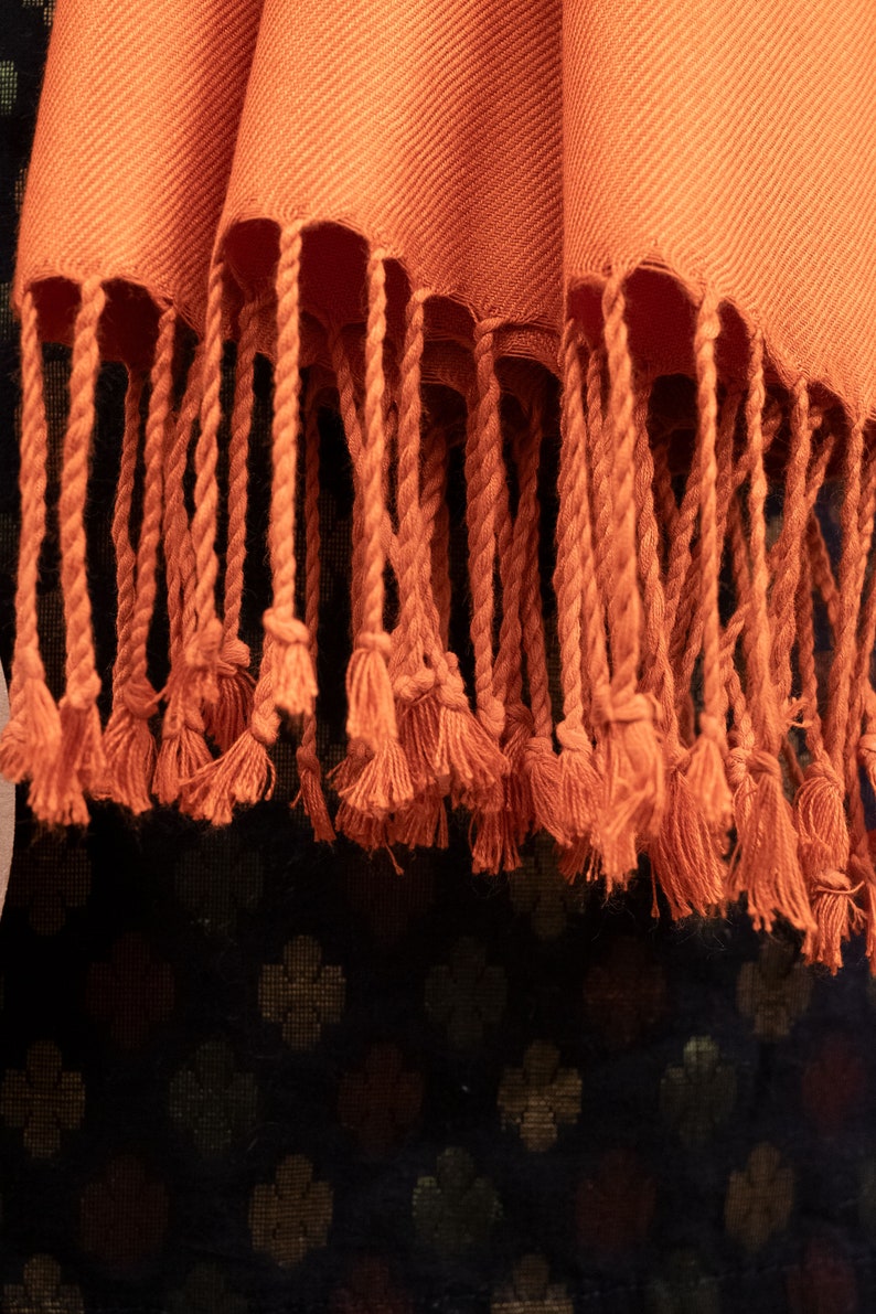 May include: A close-up of a burnt orange scarf with long fringe tassels. The scarf is made of a soft, woven fabric.