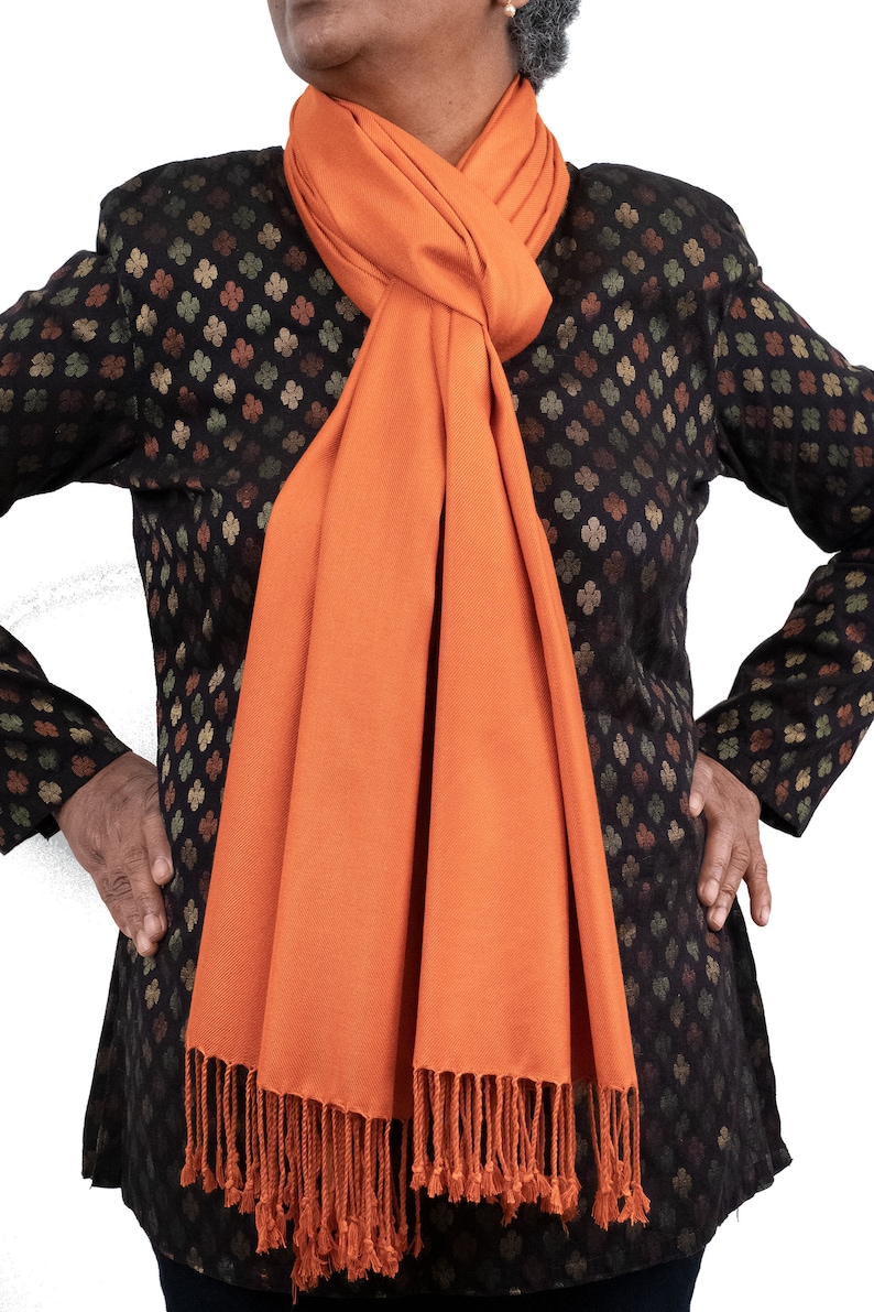 May include: A long, orange scarf with fringe on the ends. The scarf is tied around the neck in a knot.