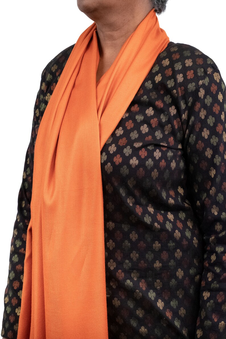 May include: A woman wearing a black and brown patterned long-sleeved shirt with an orange scarf draped around her neck.