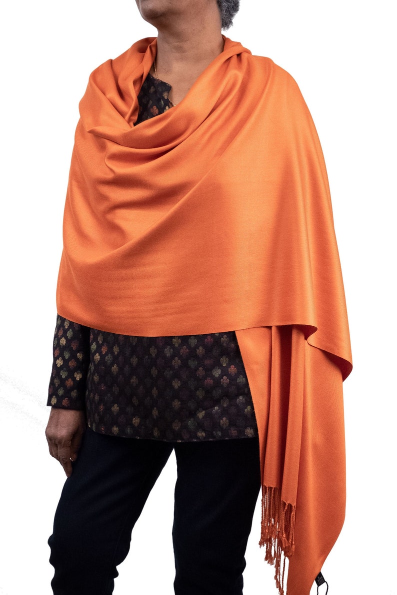 May include: A bright orange pashmina shawl with fringed edges. The shawl is draped around a person's shoulders and is made of a soft, lightweight fabric.