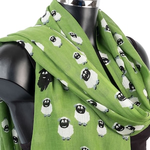 Peut inclure: Écharpe verte ornée d'un motif répété de moutons blancs avec des visages et des pattes noirs. Un mouton noir est également visible. L'écharpe est drapée sur un mannequin noir, mettant en valeur son design et sa texture.