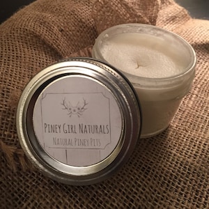 May include: A glass jar with a white lid containing a white substance. The jar has a label that reads "Piney Girl Naturals Natural Piney Pits".
