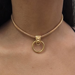 May include: Gold-toned necklace featuring a textured chain and a pendant with a lion's head design holding a large ring. The necklace is worn against a black top.