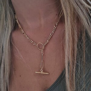 May include: A gold chain necklace with a t-bar pendant. The necklace is worn by a person with long blonde hair and a green tank top.