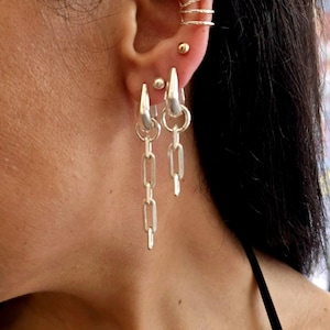 May include: Silver chain earrings with a dangling design. The earrings are paired with a silver ear cuff and a small gold stud earring. The jewellery is worn on a person's ear, with the focus on the earrings and ear cuff.