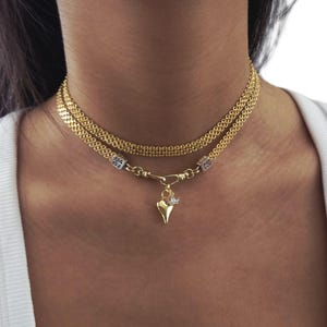 Puede incluir: Un collar dorado con dos capas de cadena. El collar tiene un colgante en forma de corazón con un pequeño dije. El collar se lleva alrededor del cuello.