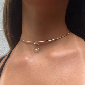 May include: A silver choker necklace with a small, round pendant. The necklace is worn around the neck. The image also shows a black tank top and a gold hoop earring.