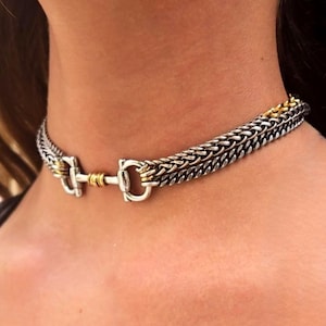 May include: A silver and gold choker necklace with a braided chain design. The necklace features a horse bit-style clasp with gold accents. The jewelry is worn around the neck.