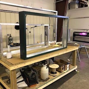 May include: A workshop scene featuring window frames in various stages of construction. A dark green frame sits on a wooden workbench, alongside other unfinished aluminum frames. Tools and equipment are visible, suggesting a manufacturing or assembly process.