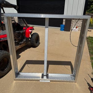 May include: A rectangular, silver-colored metal frame with a central vertical support. The frame is lying on a concrete surface, with a red ATV visible in the background. The frame appears to be a window or door frame.