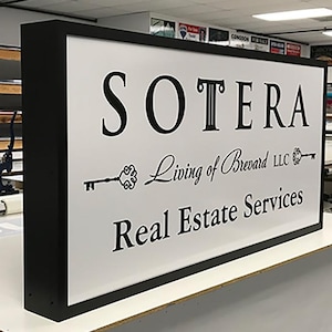May include: A rectangular sign with a black frame and a white background. The sign displays the name "SOTERA" in large, black, serif font, along with the text "Living of Brevard LLC" and "Real Estate Services" in a smaller, elegant font.