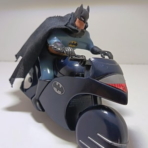 Puede incluir: Una figura de acción de Batman montada en una motocicleta azul oscuro. La figura lleva una capa de tela negra y un traje azul y gris con un emblema de murciélago amarillo. La moto tiene un logo plateado en el lateral y detalles plateados.