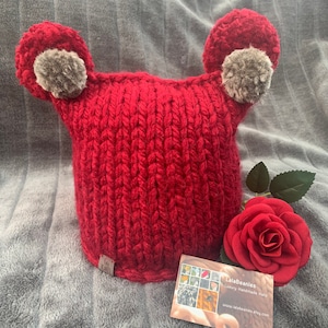 May include: Red knitted hat with grey pom-poms on the ears. The hat is handmade and has a tag that says "lalaBeanies Luxury Handmade Hats" and a website address.