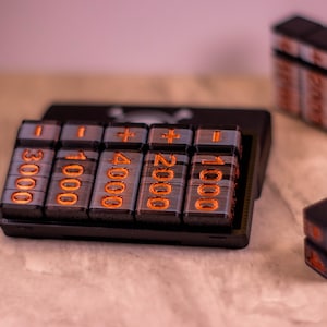 May include: A black calculator with orange illuminated numbers and symbols. The calculator has a series of buttons with numbers like 3000, 1000, 4000, 2000, and 1000. There are additional stacks of the same buttons and two dice.