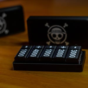 May include: Black electronic device with five buttons displaying numbers and a plus sign. Two black boxes with a skull and crossbones logo are in the background. The items are on a wooden surface.