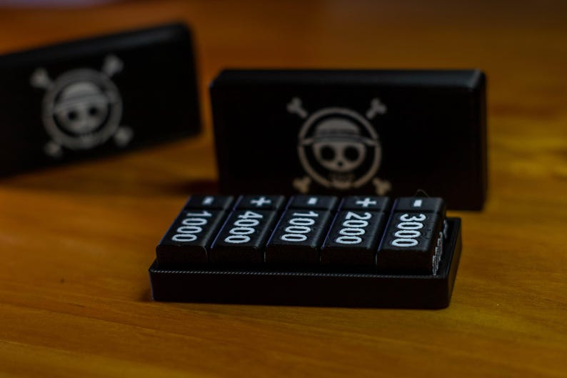 May include: Black electronic device with five buttons displaying numbers and a plus sign. Two black boxes with a skull and crossbones logo are in the background. The items are on a wooden surface.