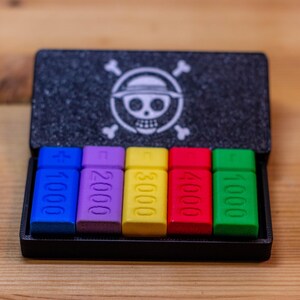 May include: A black box with a skull and crossbones logo, containing six multi-colored rectangular pieces. Each piece is labeled with a number, ranging from 1000 to 5000, in white text. The pieces are blue, purple, yellow, red, and green.