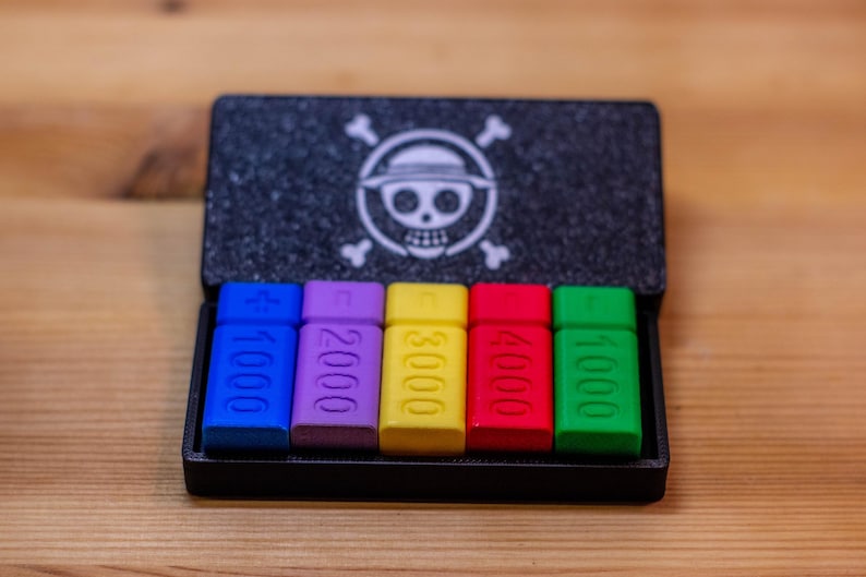 May include: A black box with a skull and crossbones logo, containing six multi-colored rectangular pieces. Each piece is labeled with a number, ranging from 1000 to 5000, in white text. The pieces are blue, purple, yellow, red, and green.