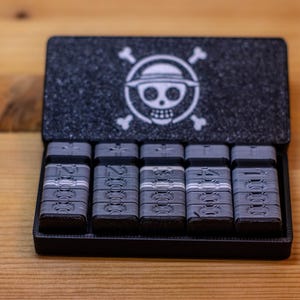 May include: A black case with a textured lid featuring a white skull and crossbones logo with a straw hat. Inside are five gray rectangular objects with white stripes and numbers. The case is open, revealing the contents.