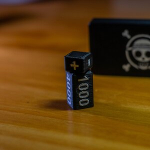 May include: Black and silver dice with the numbers 1000 and 4000, and a plus sign. A black box with a white skull and crossbones logo is in the background. The dice are on a wooden surface.