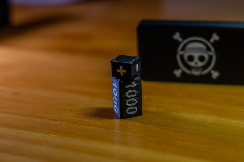 May include: Black and silver dice with the numbers 1000 and 4000, and a plus sign. A black box with a white skull and crossbones logo is in the background. The dice are on a wooden surface.