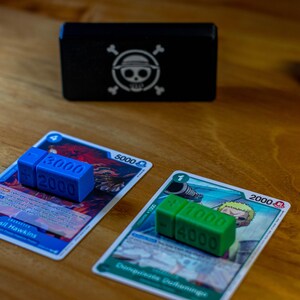 May include: A wooden table displays two trading cards with blue and green dice-shaped counters. A black box with a white skull and crossbones logo is in the background. A stack of red blocks is also visible.