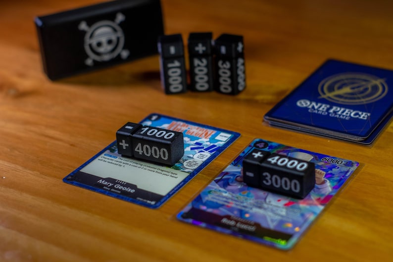 May include: Black dice with white numbers and symbols are displayed on trading cards. The dice show values like +4000 and -3000. A blue card game box and other cards are also visible. The cards have text and graphics.