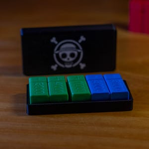 May include: A black box with a skull and crossbones logo, and a tray with green and blue rectangular pieces. The green pieces have the numbers 3000 and 4000, while the blue pieces also have the number 3000. The items are on a wooden surface.