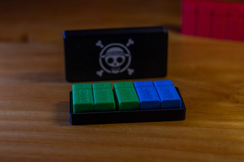 May include: A black box with a skull and crossbones logo, and a tray with green and blue rectangular pieces. The green pieces have the numbers 3000 and 4000, while the blue pieces also have the number 3000. The items are on a wooden surface.