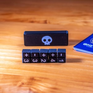 May include: A black calculator with white numbers and symbols, and a skull design. The calculator is next to a deck of blue cards with the text "ONE PIECE CARD GAME". The numbers are 1, 2, 3, 4.
