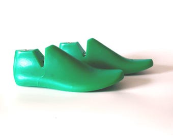 May include: Two green shoe lasts, side by side, against a white background. The shoe forms are made of a solid, smooth material. The shoe forms are a bright, solid green color.
