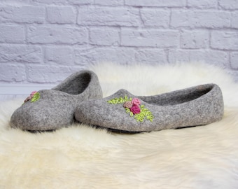 May include: A pair of gray felt slippers with embroidered pink and green floral designs. The slippers are displayed on a white, fluffy surface, with a light gray brick wall in the background. The slippers are a comfortable, cozy style.