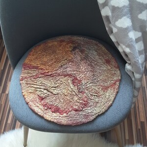 May include: A round, red, gold, and brown felted wool cushion with a textured surface. The cushion is sitting on a gray chair.