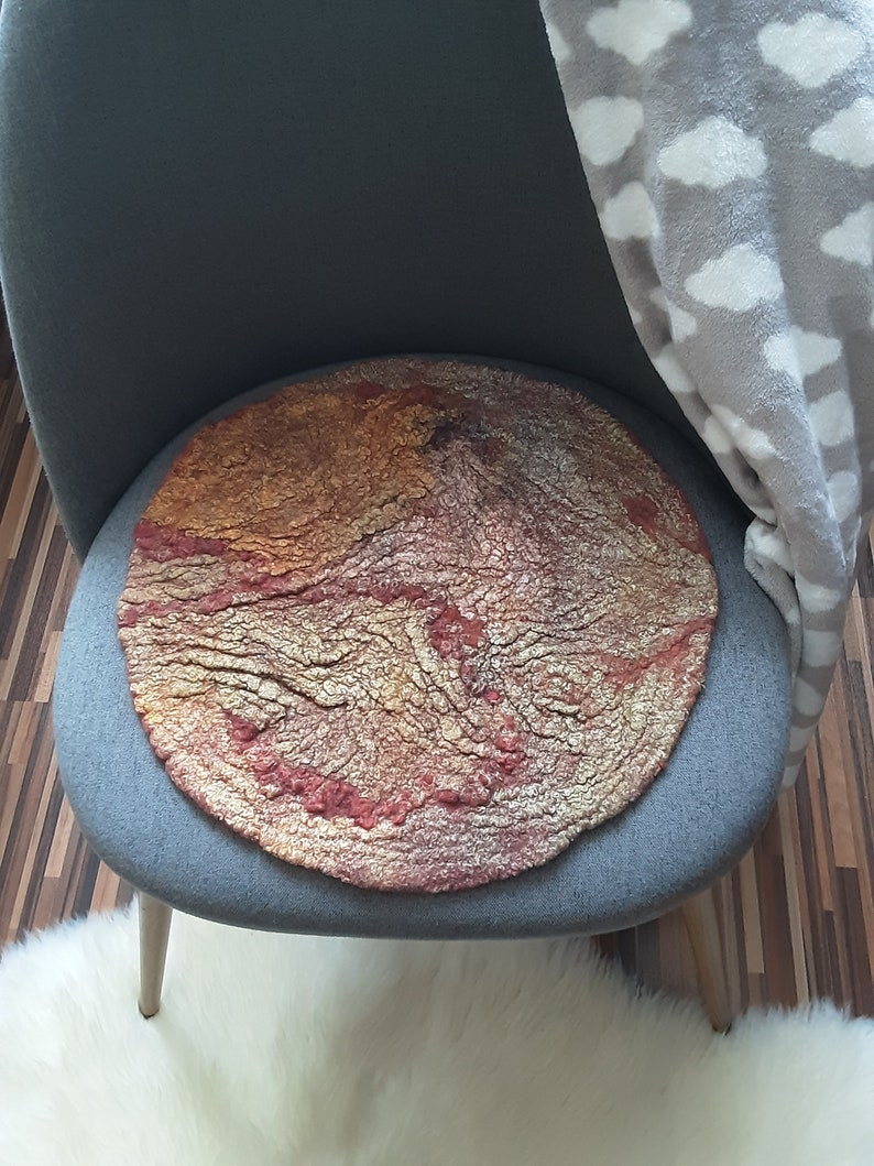 May include: A round, red, gold, and brown felted wool cushion with a textured surface. The cushion is sitting on a gray chair.