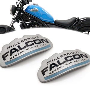 May include: A motorcycle with a light blue fuel tank and black components is displayed. Two silver-colored emblems with the text "MILLENNIUM FALCON KESSEL RUN EDITION" are in the foreground. The motorcycle has a black seat and tires.