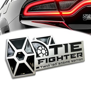 May include: A silver and black metal car emblem with a TIE fighter symbol and the text "TIE FIGHTER TWIN ION ENGINE EDITION".