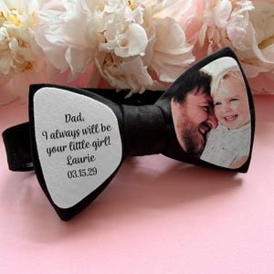 May include: A black bow tie featuring a white fabric panel on the left side with the printed text: Dad, I always will be your little girl! Laurie 03.15.29. The right side of the bow tie displays a printed photograph of an adult and a child.