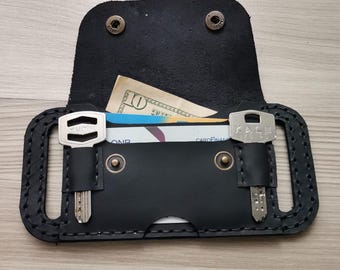 May include: Black leather key and card holder with two key slots and a snap closure. The holder contains a ten-dollar bill and a credit card. The holder is on a light wood surface.