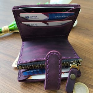Op de afbeelding: Een handgemaakte, paarse leren portemonnee met zichtbare stiksels. De portemonnee heeft meerdere kaartsleuven, een vak met ritssluiting en een drukknoopsluiting. De portemonnee is open en toont creditcards en wat contant geld.