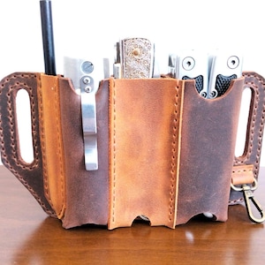 Pode incluir: Um organizador multi-ferramentas de couro castanho artesanal. Possui vários bolsos, um porta-canetas e um clipe de metal. Contém várias ferramentas, incluindo uma faca e uma multi-ferramenta. O design inclui bordas costuradas e uma presilha para cinto.