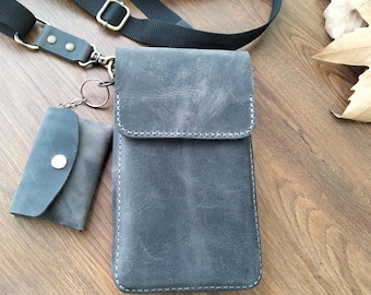 May include: A gray leather crossbody bag with a matching small wallet attached by a keyring. The bag has a flap closure and white stitching. A black adjustable strap is attached to the bag. The bag is approximately 7 inches tall.