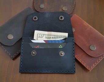 May include: A collection of handmade leather wallets in various shades of brown and black. One wallet is open, revealing a ten-dollar bill and several cards. Each wallet features a snap closure and visible stitching.