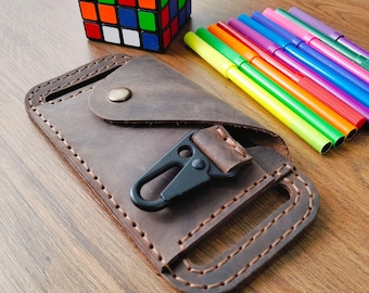 May include: A brown leather key holder with a black carabiner clip. The key holder is rectangular with a button closure and stitched edges. A colorful Rubik's Cube and a set of colorful markers are in the background.