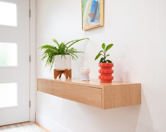 Op de afbeelding: Een zwevend houten plank met een lade, een pot met een varen, een kleine witte keramische pot met een geometrisch ontwerp en een rode keramische pot met een gestapeld ontwerp.