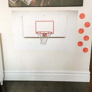 May include: A white basketball hoop with a red rim and a black net is mounted on a white wall. There are several orange basketballs with black outlines on the wall around the hoop.